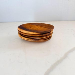 May include: A stack of five small, round wooden plates. The plates are a warm brown color with visible wood grain. The plates are stacked on top of each other, showcasing their circular shape and natural wood texture.