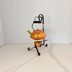 May include: A copper-colored tea kettle with a matching burner sits on a black metal stand. The kettle has a rounded body, a spout, and a handle. The stand has decorative scrollwork and a circular base. The setup is designed for heating tea.