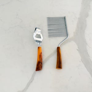 May include: Two kitchen tools on a white marble surface. One is a stainless steel cheese grater with an orange and brown handle. The other is a cake leveler with a wooden handle. Both tools are designed for food preparation.