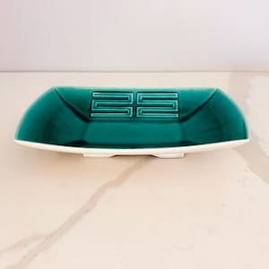 May include: A rectangular ceramic dish with a deep teal glazed interior and a white exterior. The center of the dish features a raised, geometric Greek key-style pattern. The dish has slightly curved sides and rests on a small base.