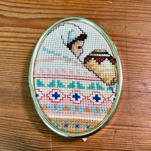 May include: Oval framed cross-stitch embroidery depicting a figure in a headscarf and a decorative vessel. The embroidery uses white, pink, blue, and brown. The frame is gold-coloured and the background is a wooden surface.
