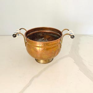 May include: A round copper bowl with a polished finish, featuring two curved metal handles on opposite sides. Each handle is accented with a white ceramic insert decorated with a blue floral pattern. The bowl sits on a circular pedestal base.