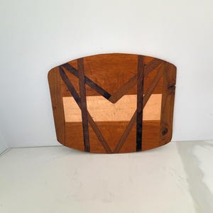 May include: Wooden decorative wall art with a geometric design. The piece features a combination of light and dark wood tones, with a central light-colored section and dark wood accents. The overall shape is a rounded rectangle.