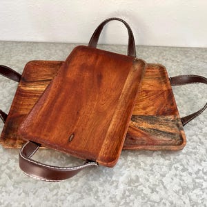 May include: Two rectangular wooden serving trays with brown leather handles and white stitching. The trays have a rich, reddish-brown color with visible wood grain. One tray is partially covering the other.