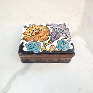 May include: A square ceramic trinket box with a floral design. The lid features a large orange and yellow flower and a purple flower with blue leaves. The box has a dark brown base with a wavy black border.
