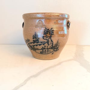 May include: A tan stoneware crock featuring a dark blue hand-painted illustration of a dragon-like creature. The cylindrical vessel has a thick rim, two small side handles, and a stamped maker's mark located near the top edge of the exterior.