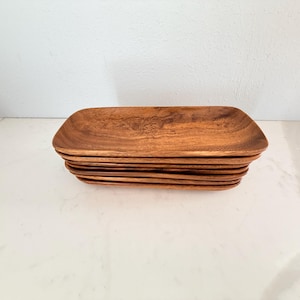 Pode incluir: Uma pilha de bandejas retangulares de madeira. As bandejas são feitas de madeira marrom clara com padrões de grãos visíveis. As bordas são ligeiramente curvas e as bandejas são empilhadas ordenadamente umas sobre as outras.