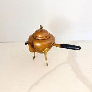 May include: A copper teapot with a rounded body, a matching lid with a small knob, and a curved spout. The teapot is supported by three slender brass legs and features a long, straight handle with a black finish and visible wear.