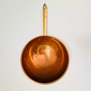 May include: A round copper bowl with a long, straight brass handle attached to the top rim. The interior of the bowl is polished, showing a reflective metallic surface. Three small rivets are visible where the handle connects to the bowl.
