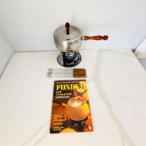 May include: A stainless steel fondue pot with a wooden handle and lid, a black metal stand, six fondue forks with wooden handles, and a Better Homes and Gardens Fondue and Tabletop Cooking cookbook with a yellow and black cover design.