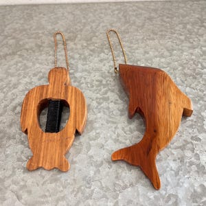 May include: Two wooden ornaments: a turtle and a shark. The turtle has a black strap through its center. Both ornaments are made of light brown wood and have gold-colored chain loops for hanging.