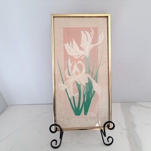 May include: A framed art print featuring a floral design with white flowers, green stems, and a pink background. The print is in a gold-colored frame and displayed on a black metal easel. The artwork has a vintage aesthetic.