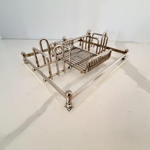 May include: A silver-toned metal toast rack with a rectangular wire basket and vertical dividers. The rack sits on a clear, rectangular base with rounded corners and decorative ball accents.