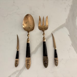 May include: A set of four antique serving utensils: a spoon, fork, and two knives. The utensils have brass heads, black handles, and decorative brass accents. The spoon and fork have a twisted design on the handles.