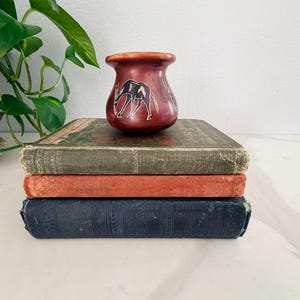 Puede incluir: Una pila de tres libros antiguos con cubiertas desgastadas, coronada por un pequeño jarrón marrón rojizo. El jarrón presenta un diseño blanco grabado de dos jirafas. Los libros son en tonos verdes, naranjas y azul marino.