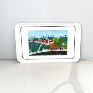 May include: A rectangular white tray with a colorful folk art painting of a village scene. The painting depicts a church, houses, and people. The tray has a black border and is set against a white background.