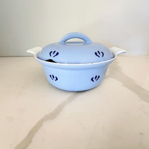 May include: A light blue ceramic casserole dish with a matching lid and two side handles. The exterior of the bowl and the lid are decorated with a repeating pattern of small, dark blue, three-petaled floral motifs arranged in a horizontal row.