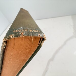 Puede incluir: Un libro antiguo con una cubierta verde desgastada y letras doradas que dicen "WISE & CO". Las p&aacute;ginas del libro est&aacute;n envejecidas y tienen un tono marr&oacute;n c&aacute;lido. La cubierta muestra signos de desgaste, lo que aumenta su atractivo vintage.