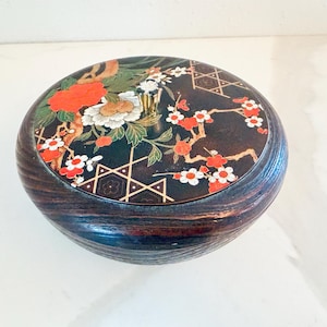 May include: A round, wooden box with a dark finish and a decorative lid. The lid features a black background with floral and geometric designs in red, white, and gold. A small, gold handle is centered on the lid.