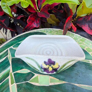 May include: A speckled, off-white ceramic soap dish with a floral design. The dish has a curved shape with a purple and yellow flower painted on the front. The dish is sitting on a green mosaic table.