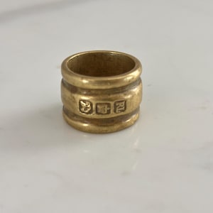 Puede incluir: Un anillo dorado con forma cilíndrica. El anillo tiene tres bandas horizontales y está marcado con tres pequeños símbolos. El anillo está sobre una superficie blanca.