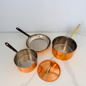 May include: A set of copper cookware consisting of two saucepans with metallic handles, one shallow frying pan with a dark handle, and a circular copper lid with a central handle. The interior of each piece is silver-toned metal.