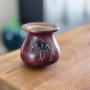 May include: A small, round, maroon-colored vase with a black etched design of an antelope. The vase has a wide opening and sits on a wooden surface. The background is blurred.