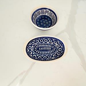 May include: A round ceramic bowl and an oval ceramic dish, both featuring a cream-colored base with intricate dark blue patterns. The bowl displays a geometric Greek key border, while the oval dish is decorated with concentric circles and dots.