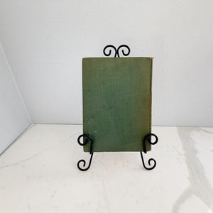 Puede incluir: Un libro antiguo con una cubierta verde descolorida se exhibe en un soporte de hierro forjado negro. El soporte tiene adornos decorativos y sostiene el libro en posici&oacute;n vertical. El fondo es una pared blanca y una encimera blanca.