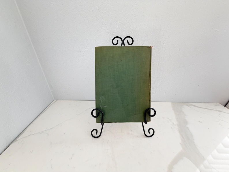 Puede incluir: Un libro antiguo con una cubierta verde descolorida se exhibe en un soporte de hierro forjado negro. El soporte tiene adornos decorativos y sostiene el libro en posici&oacute;n vertical. El fondo es una pared blanca y una encimera blanca.