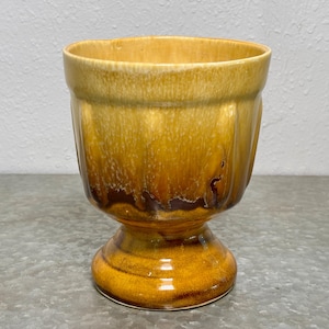 May include: A vintage, pedestal-style planter with a glossy, glazed finish. The ceramic planter features a warm color palette of golden yellow, brown, and amber tones. The exterior has vertical ribbed detailing. The planter is suitable for indoor or outdoor use.