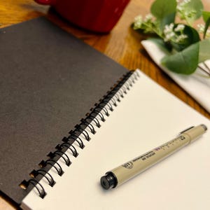 Puede incluir: Un cuaderno abierto con espiral, con páginas en blanco y negro, y un bolígrafo beige con tinta negra. Una taza roja y vegetación en el fondo. El bolígrafo está etiquetado como "ARCHIVAL INK".