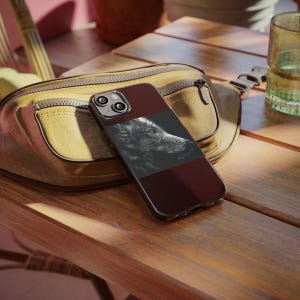 Op de afbeelding: Een smartphonehoesje met een donkerrood en zwart ontwerp met een zwart-wit wolf illustratie. De hoes ligt op een houten oppervlak naast een gele heuptas.