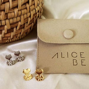 Pode incluir: Dois pares de elevadores de brincos em forma de coração, um prateado e outro dourado, posicionados ao lado de uma bolsa de camurça bege com fecho de botão e o texto ALICE BE impresso a preto, junto a um cesto de vime entrançado.