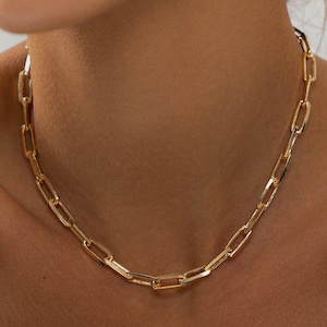 May include: A gold-toned chain necklace featuring elongated, rectangular links connected in a repeating pattern. The necklace rests around the neck, showcasing a polished, metallic finish with uniform, open-style links that create a continuous, structured design.