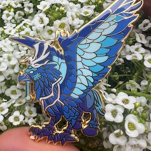 May include: A detailed enamel pin featuring a blue griffin design with gold accents. The griffin has a fierce expression and is set against a backdrop of white flowers. The pin is a vibrant accessory.