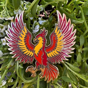 Könnte beinhalten: Ein farbenfroher Phönix-Ansteckpin mit detailliertem Design. Der Pin hat ein rot-, gelb- und oranges Farbschema mit goldenen Konturen. Der Phönix ist mit ausgebreiteten Flügeln dargestellt, seine Krallen sind sichtbar. Der Pin ist vor grünem Laub platziert.