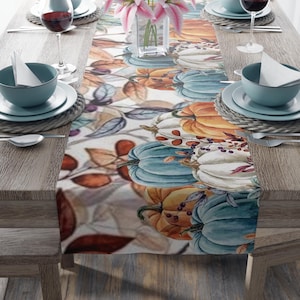 Op de afbeelding: Een tafelloper met een aquarelontwerp van pompoenen in oranje, blauw en wit, met herfstbladeren en bessen. De loper ligt op een houten tafel, met couverts en wijnglazen.
