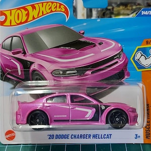 以下が含まれることがあります： ピンク色の2020年型ダッジ・チャージャー・ヘルキャットのホットウィールダイキャストモデル。おもちゃの車は黒いアクセントとホイールがあり、青とオレンジのブリスターパックに入っています。パッケージには「Hot Wheels」と「Muscle Mania」の文字があります。