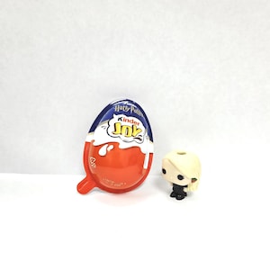 May include: A Kinder Joy egg in orange, white, and blue, featuring "Harry Potter" and "Kinder Joy" branding. Beside it is a small figurine of a character with blonde hair and a black outfit.