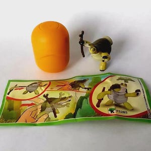 Op de afbeelding: Een oranje plastic ei naast een klein Kung Fu Panda speelgoedfiguur van Meester Oogway. De figuur is groen, geel en wit en houdt een bruine staf vast. Er is ook een groen en wit papier met illustraties van het personage.