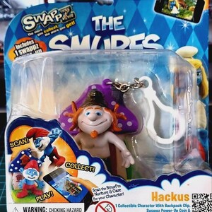 Może przedstawiać: Zapakowana zabawka przedstawiająca postać podobną do Smerfa o imieniu Hackus z fioletowym kapeluszem z grzyba i białym klipsem. Opakowanie zawiera tekst "The Smurfs" i "Swapz", a także kod QR oraz słowa "Collect!" i "Play!"