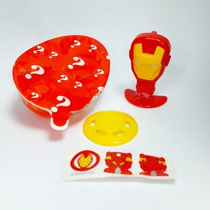 May include: A collection of Marvel Iron Man themed toys and accessories. Includes a red and yellow Iron Man head, a yellow pacifier, a sticker sheet with Iron Man graphics, and a red container with white question marks.