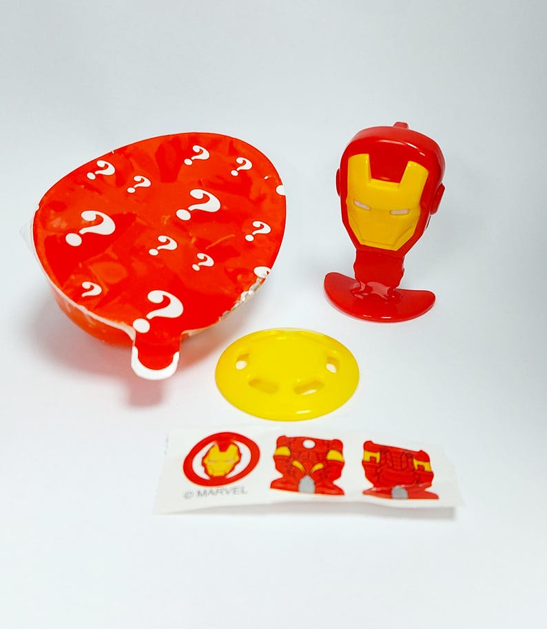 May include: A collection of Marvel Iron Man themed toys and accessories. Includes a red and yellow Iron Man head, a yellow pacifier, a sticker sheet with Iron Man graphics, and a red container with white question marks.