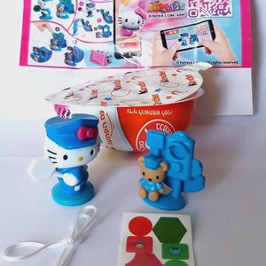 May include: A Kinder Surprise egg with a Hello Kitty figurine and a small bear figurine, both in blue and white. Includes a sticker sheet and a white string. The packaging features a QR code and the text "KINDER.COM/APP".