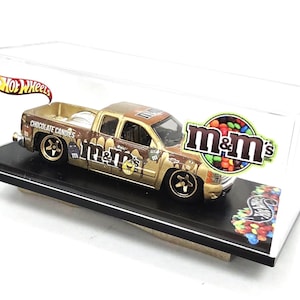 May include: A Hot Wheels die-cast pickup truck featuring an M&M's chocolate candies theme with brown and gold paint, M&M's character graphics, and the text "CHOCOLATE CANDIES" on the side, enclosed in a clear plastic display case with a black base.