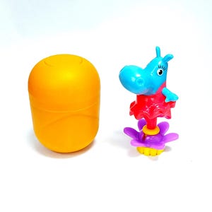 Puede incluir: Una figura de hipopótamo azul con un vestido rojo y una base de flor morada se encuentra junto a un huevo de plástico naranja. El hipopótamo tiene una gran cabeza azul, orejas pequeñas y un cuello rojo.