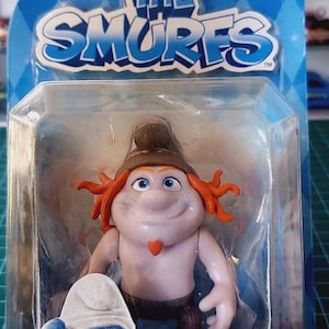 Może przedstawiać: Figurka Smerfów w przezroczystym plastikowym opakowaniu. Figurka przedstawia postać z rudymi włosami, brązowym kapeluszem i małą czerwoną brodą. Opakowanie zawiera tekst "The Smurfs" i "Movie Grab 'Ems".