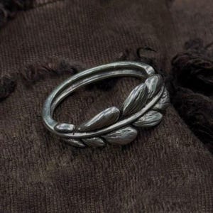 May include: A silver ring with a leaf motif. The ring has a double band and a detailed leaf design on one side. The ring is displayed on a dark brown, textured surface.