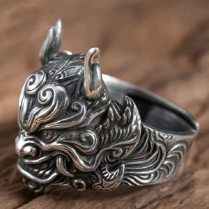 May include: A silver ring with a detailed demon mask design. The ring features intricate carvings, including horns and swirling patterns, with a dark, oxidised finish. A striking piece of jewellery.